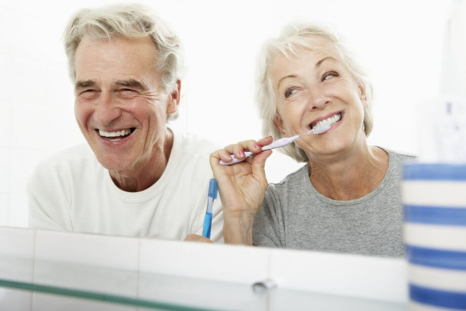 Photograph of an older couple in a bathroom. The woman is brushing her teeth & the man is smiling while looking in the mirror