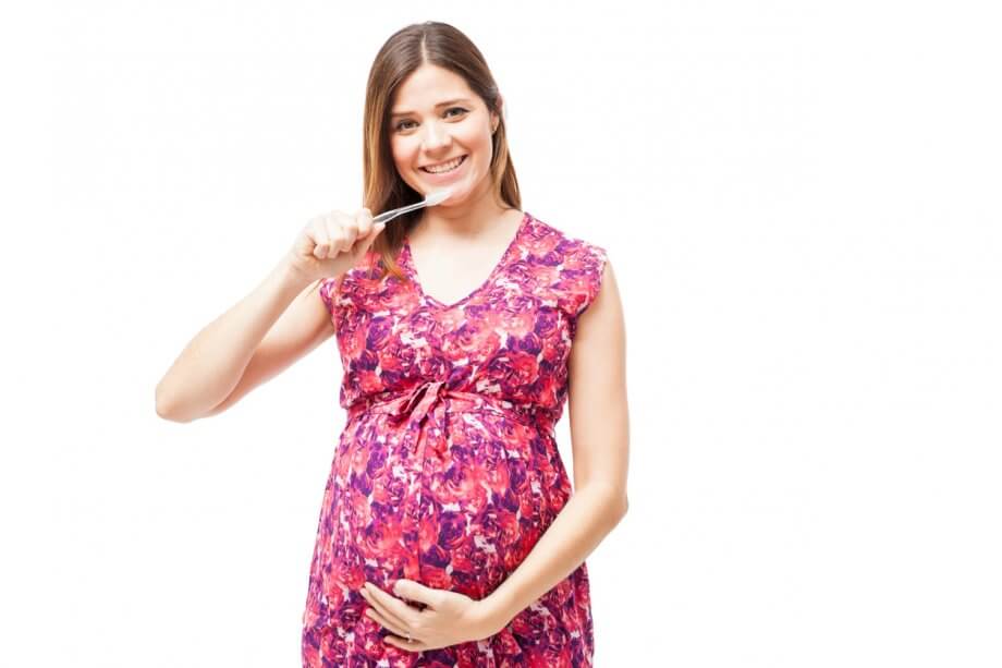 Pregnant woman with straight brown hair smiling while holding her belly with one arm and a toothbrush in the other.