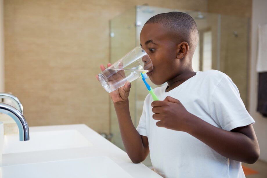 Young boy in front of a bathroom sink with a toothbrush in his hand as he takes a sip from a glass of water.